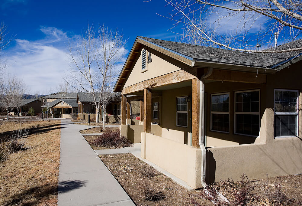 Single-story cottage home with reclaimed-timber porch posts and a walkway to neighboring homes at La Tierra in Ruidoso, New Mexico